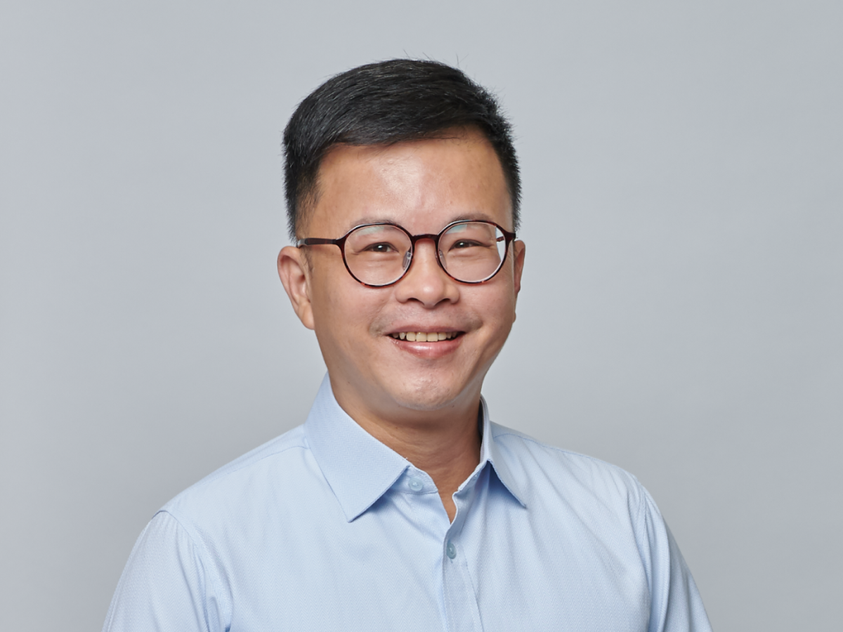 Mr Mak Mun Whai in glasses and a light blue shirt smiling with hands loosely clasped against a plain grey background